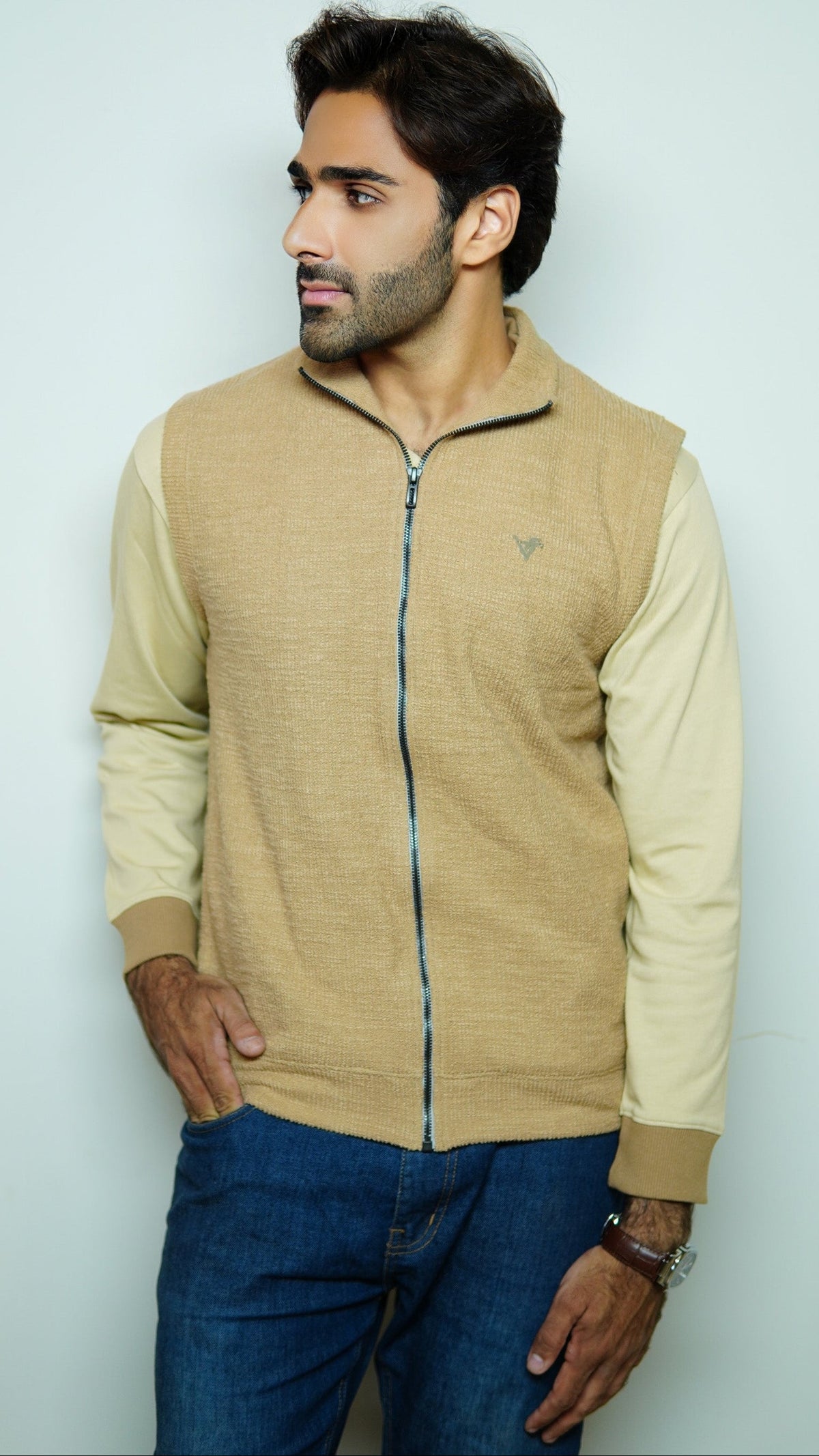 Man wearing a beige zip-up jacket and blue jeans against a light gray background